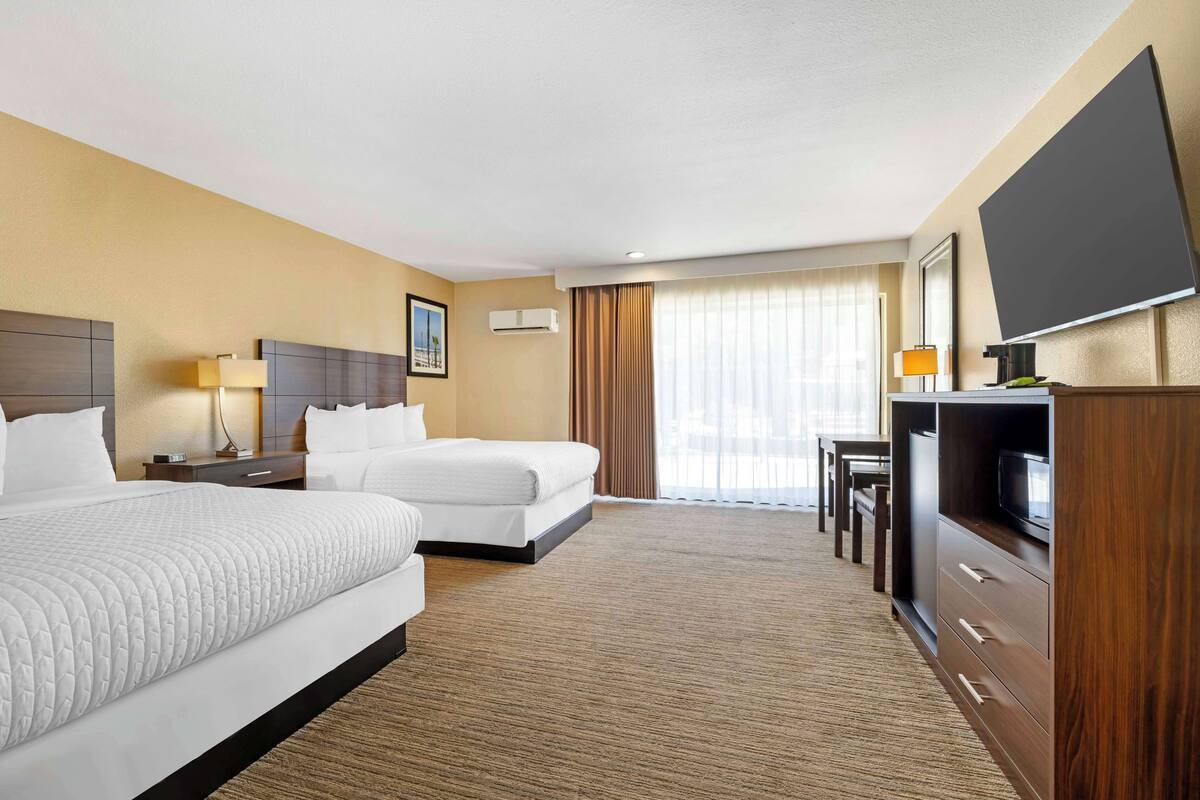 A hotel room with two beds, a TV, a desk with a chair, and a large window with curtains.