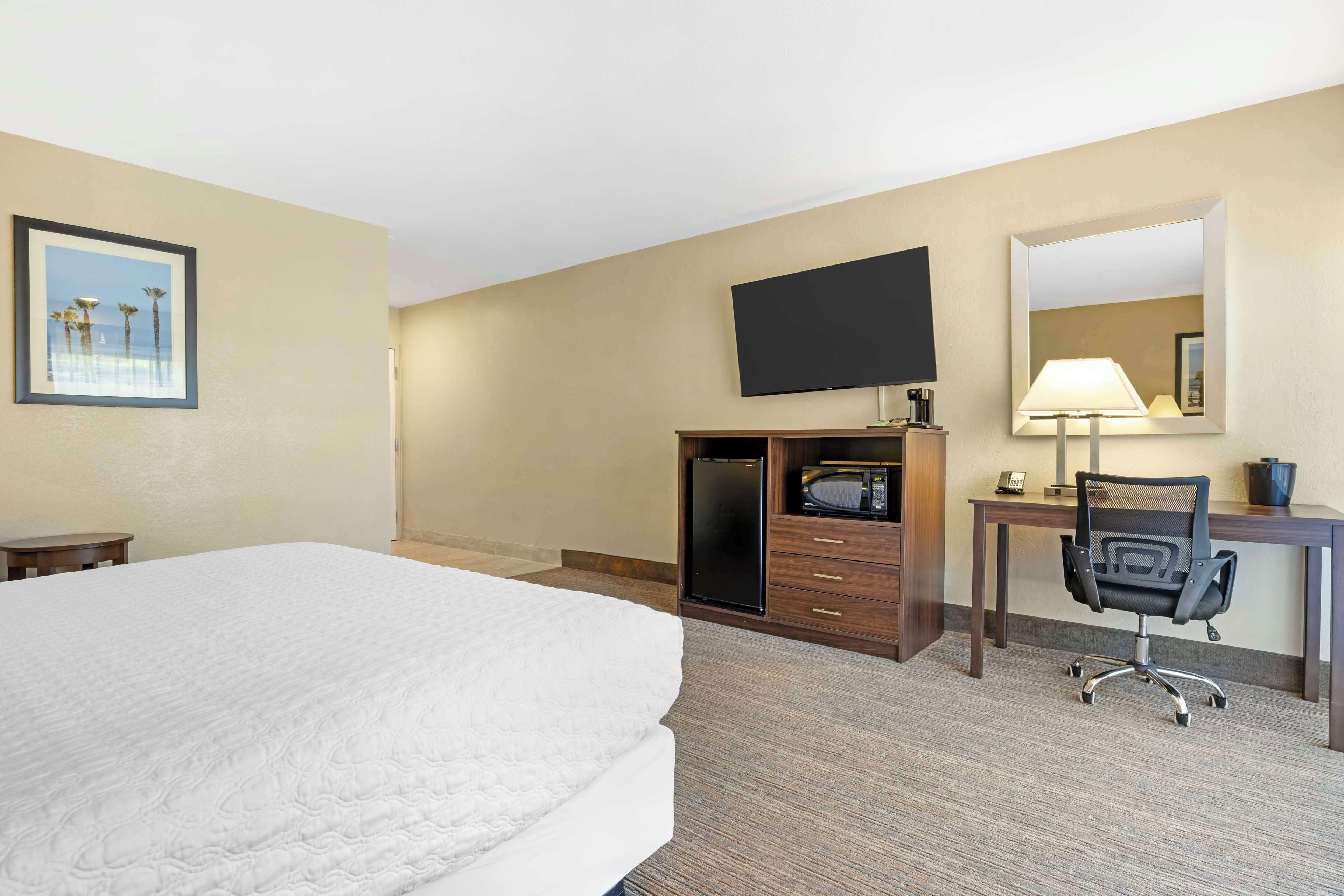 A hotel room with a large bed, a desk with a chair, a flat-screen TV, a desk lamp, a mirror, and a framed picture on the wall.
