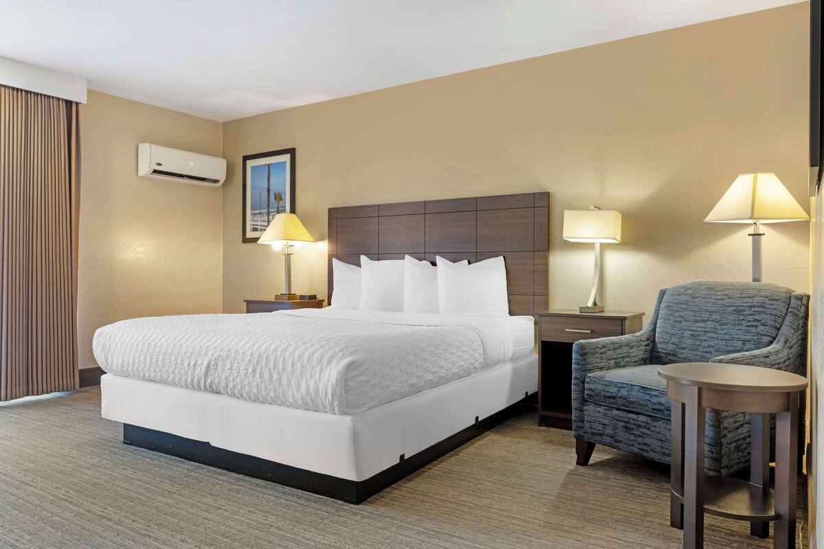 A hotel room with a large bed, two bedside tables with lamps, a chair, and an air conditioning unit on the wall.