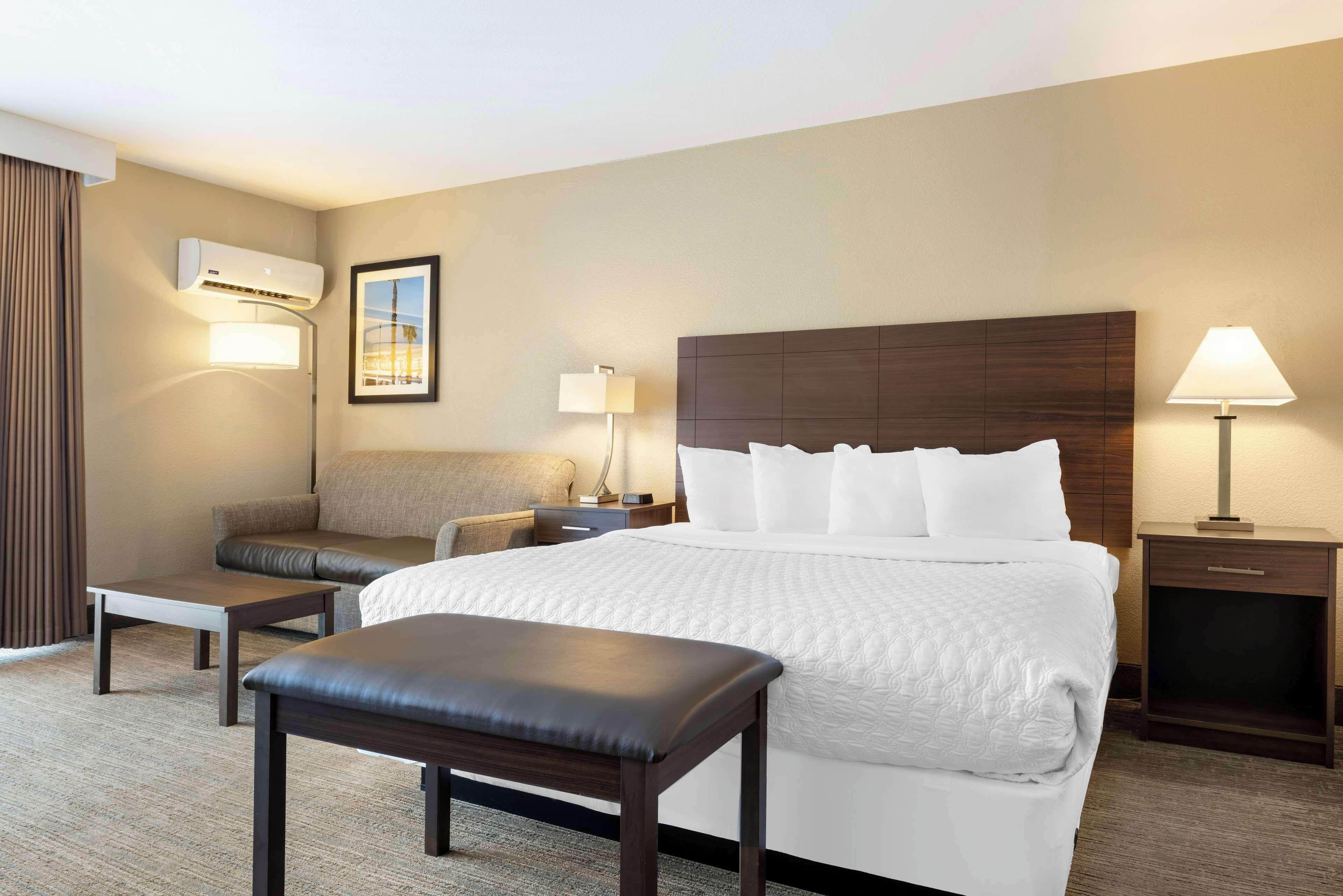 Una habitación de hotel con una cama grande, un sofá, un banco, una mesita de noche con lámpara y un equipo de aire acondicionado.
