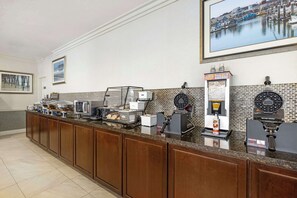 A breakfast bar with coffee machines, a juice dispenser, and a buffet with various food items.