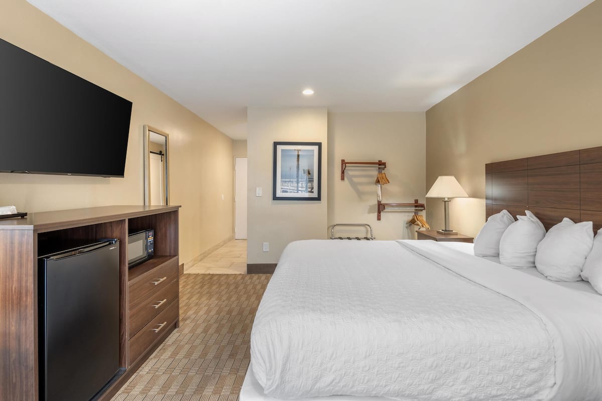 A hotel room with a bed, a TV mounted on the wall, a desk with a microwave, and a bedside table with a lamp.