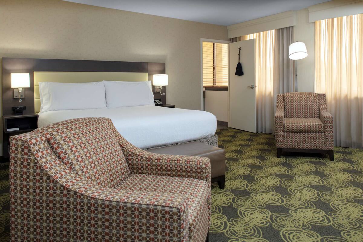 Ein Hotelzimmer mit einem großen Bett, zwei Nachttischen mit Lampen, einem gemusterten Sessel, einem kleinen Hocker und einem teppichartigen Bodenbelag.