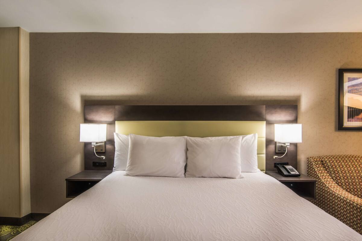 Ein Hotelzimmer mit einem großen Bett, zwei Nachttischlampen, einem Telefon und einem gemusterten Sessel.