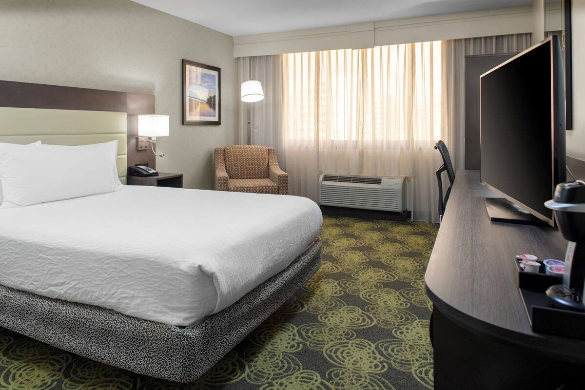 Ein Hotelzimmer mit einem großen Bett, einem Schreibtisch mit Fernseher, einem Sessel und einer Klimaanlage.