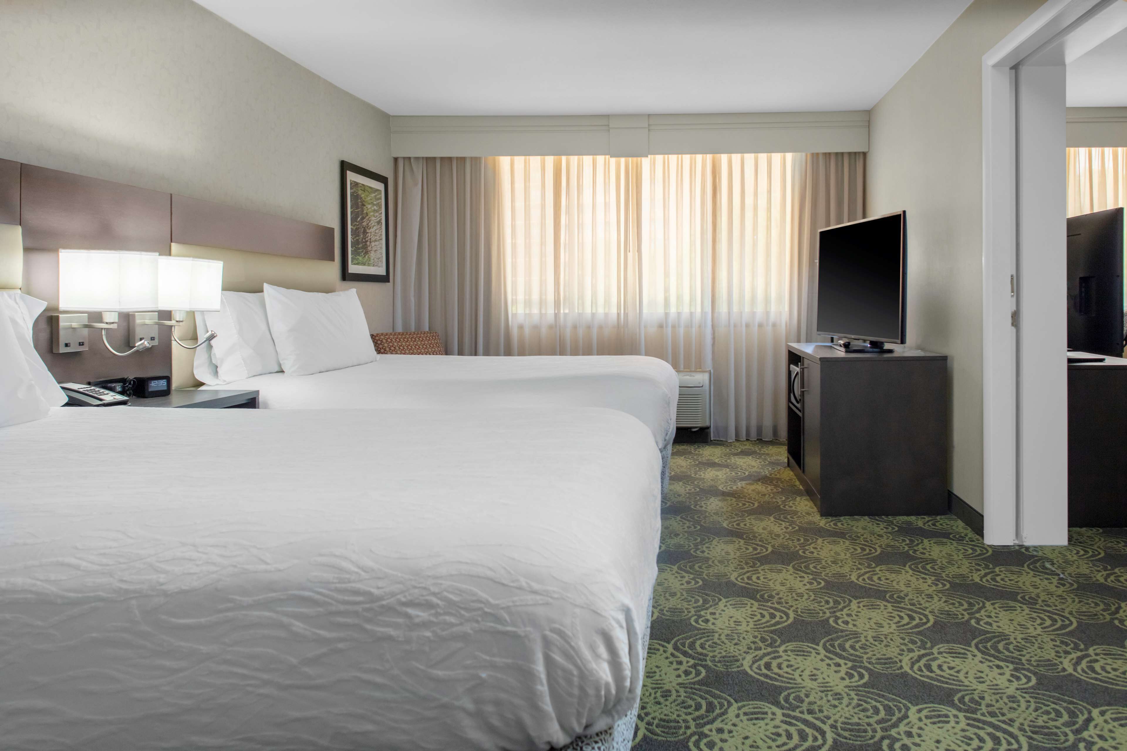 A hotel room with two beds, a TV, a desk, and a large window with curtains.
