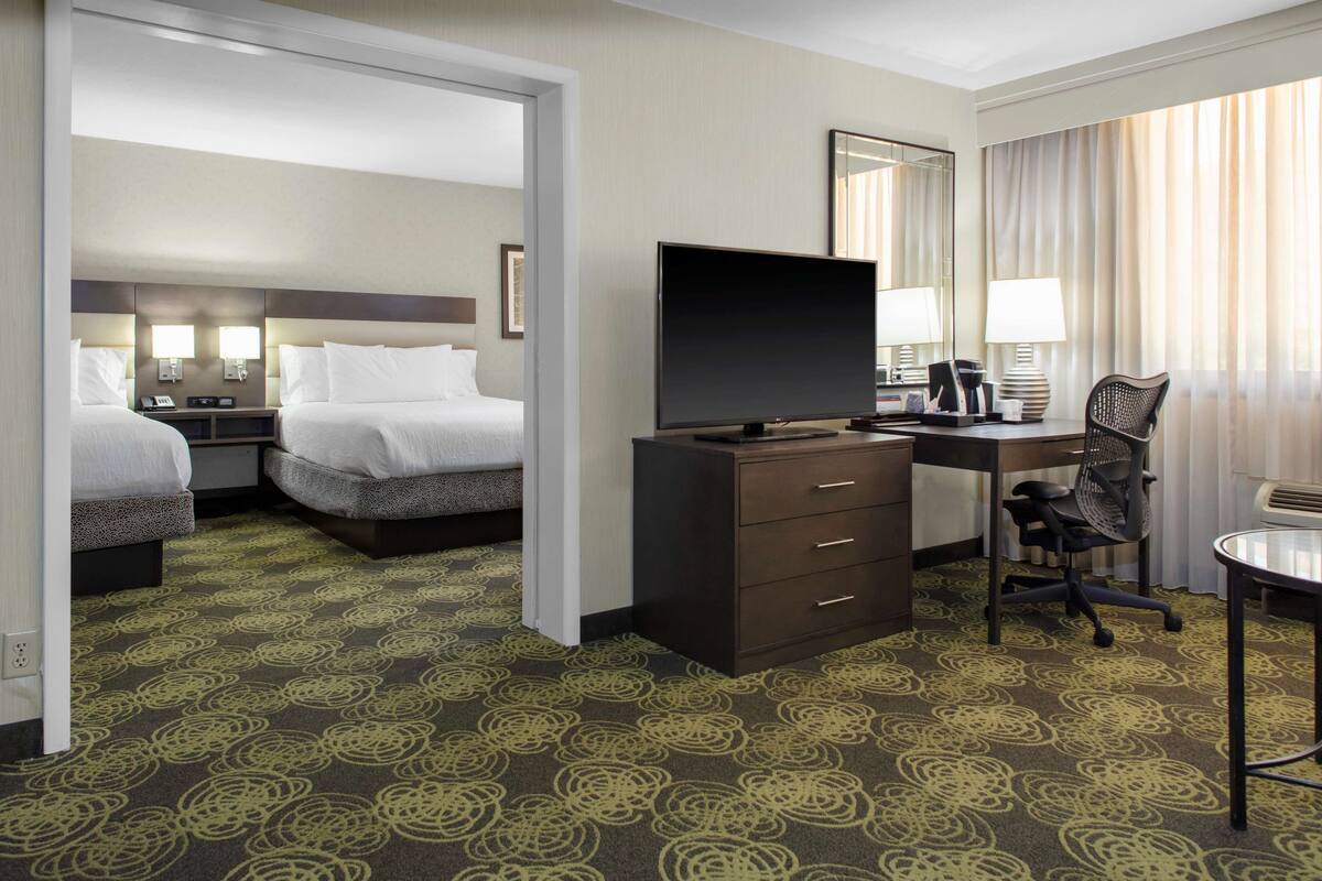 Ein Hotelzimmer mit zwei Betten, einem Schreibtisch mit Fernseher, einem Sessel und einer Schreibtischlampe.