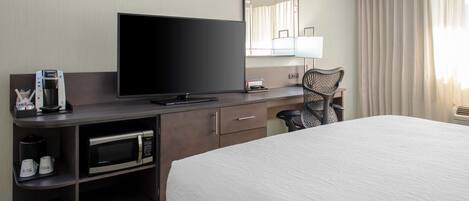 Habitación de hotel con una cama grande, un televisor de pantalla plana, cafetera, microondas, una silla y un espejo amplio.