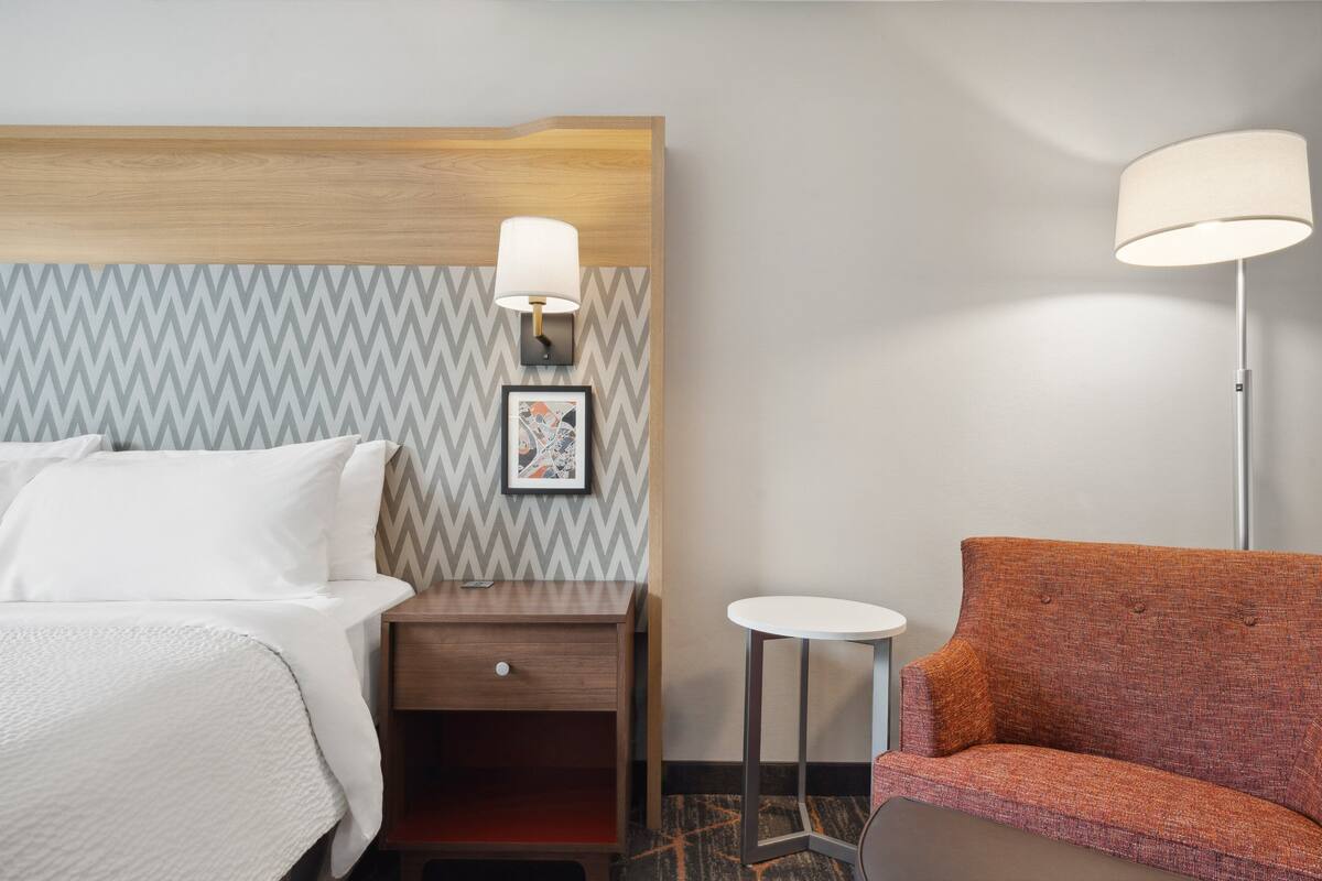 A hotel room with a bed, a nightstand, a lamp, a small table, and a sofa.
