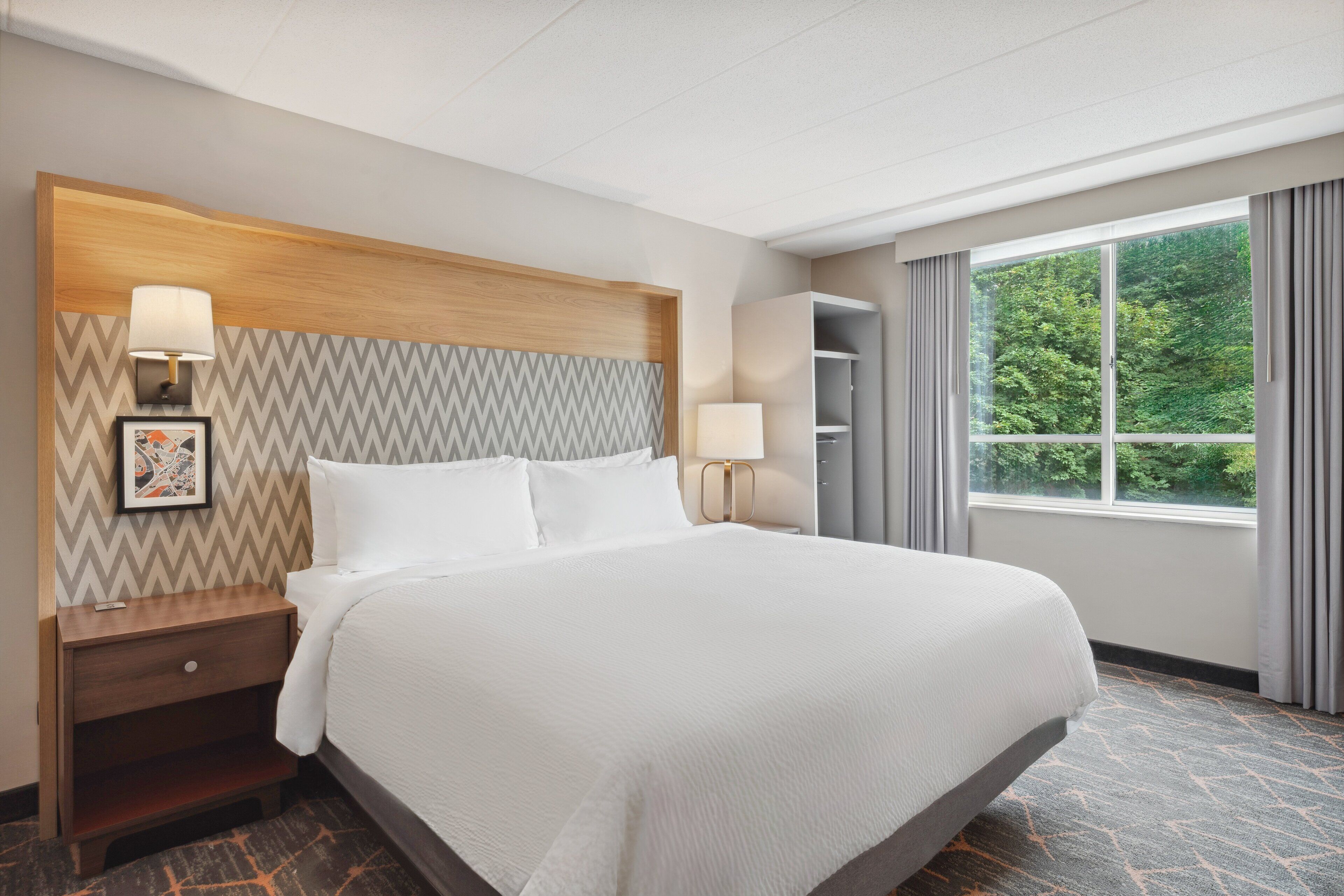 A hotel room with a large bed, a wooden headboard, a nightstand, a lamp, a picture frame, a wardrobe, and a window with curtains.
