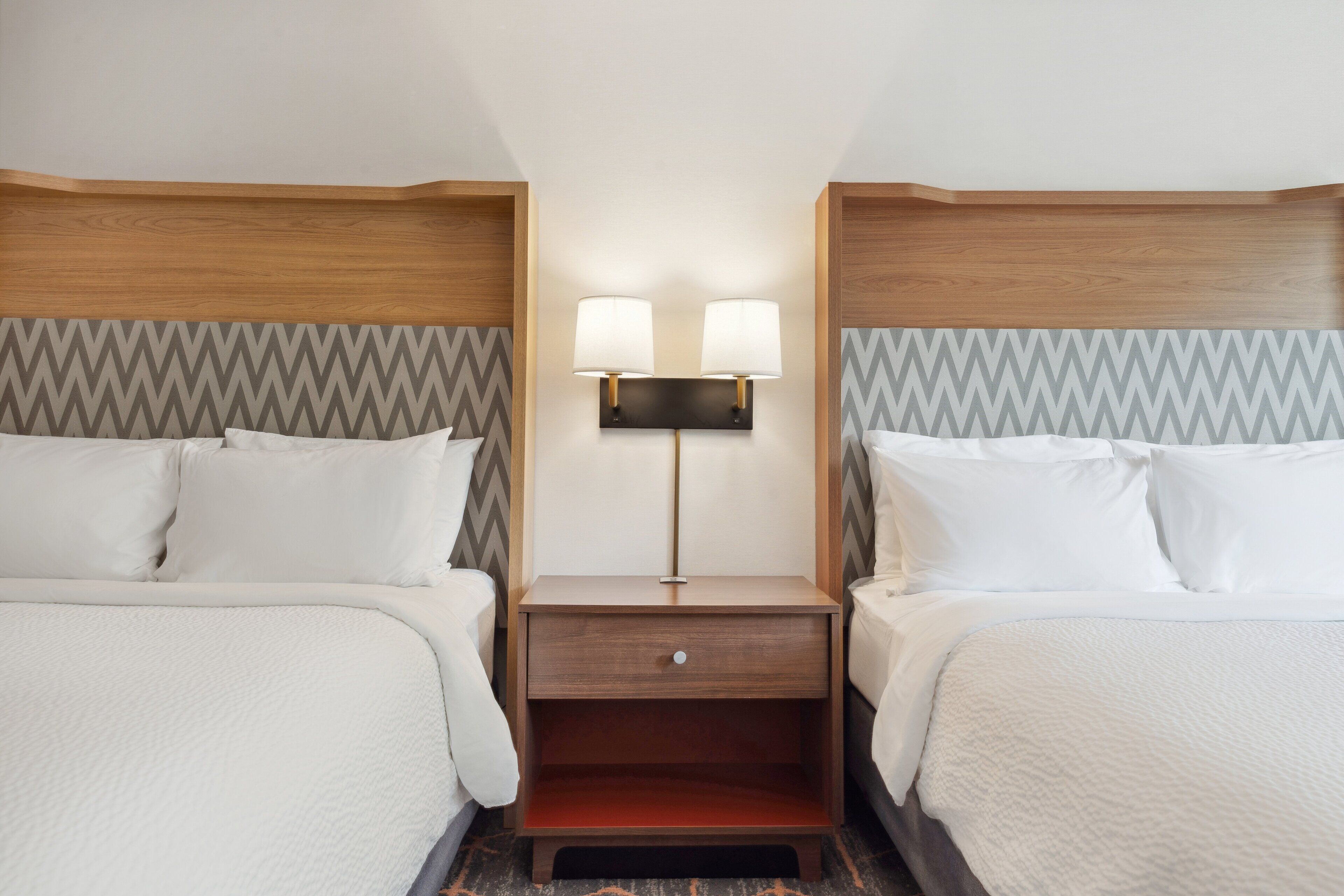 A hotel room with two beds, a nightstand with a lamp, and a chevron-patterned wall.