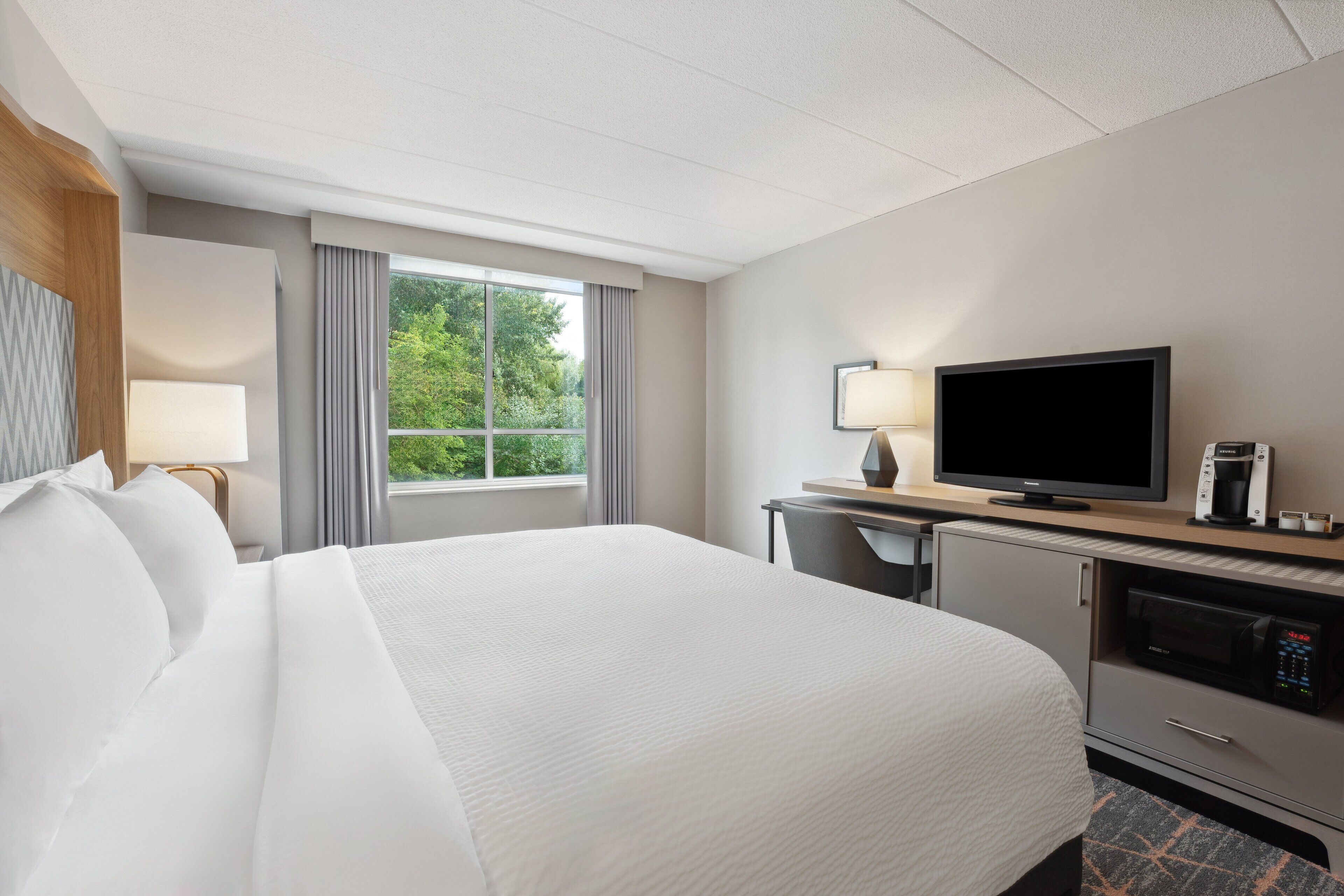 Une chambre d’hôtel avec un grand lit, une télévision, un bureau avec une lampe et une fenêtre avec des rideaux.
