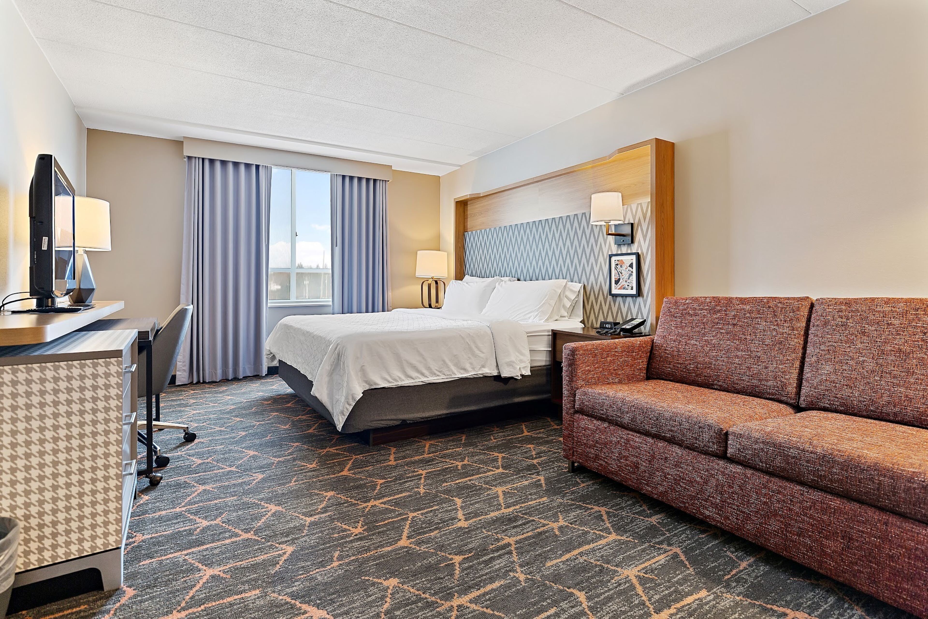 A hotel room with a bed, a desk with a computer, a sofa, and a window with curtains.