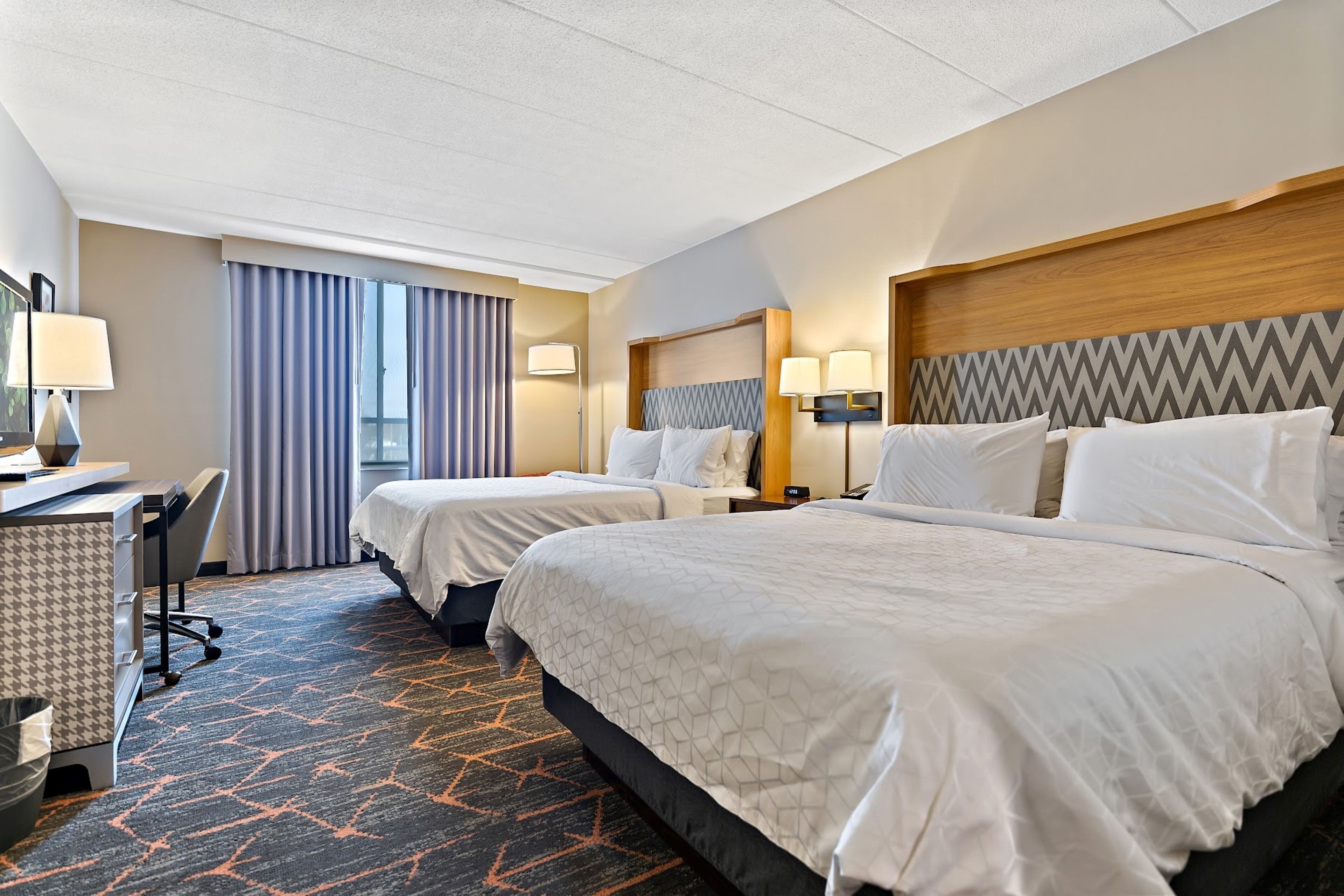 A hotel room with two beds, a desk, a chair, and a large window with curtains.