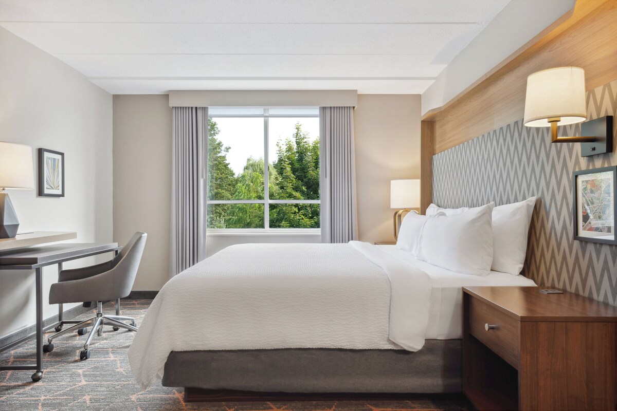 A hotel room with a bed, desk, chair, and a large window with curtains.