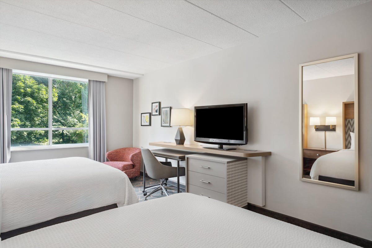A hotel room with two beds, a desk with a TV, a chair, a mirror, and a window with curtains.