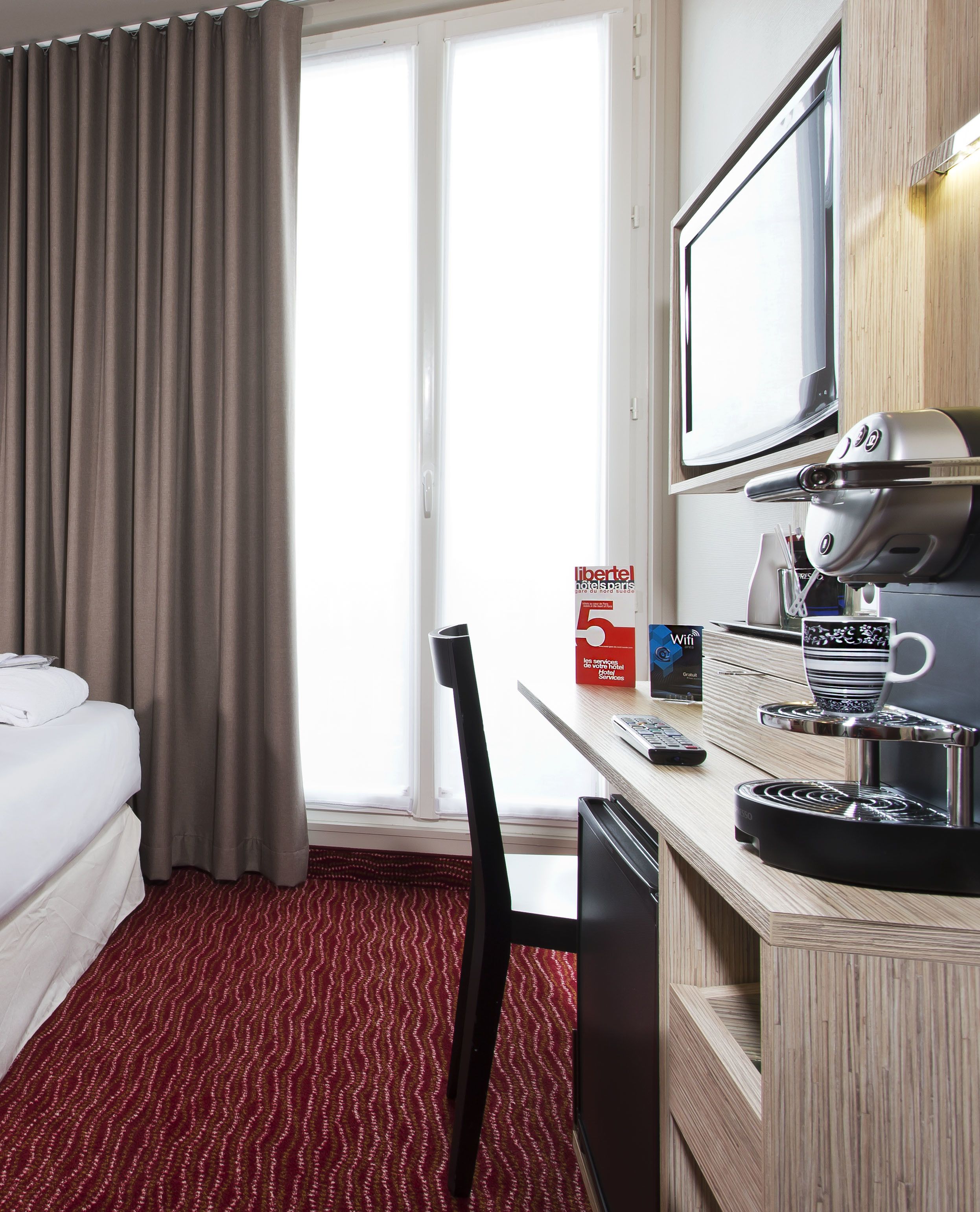A hotel room with a bed, a desk with a computer, a coffee machine, a TV, and a window with curtains.