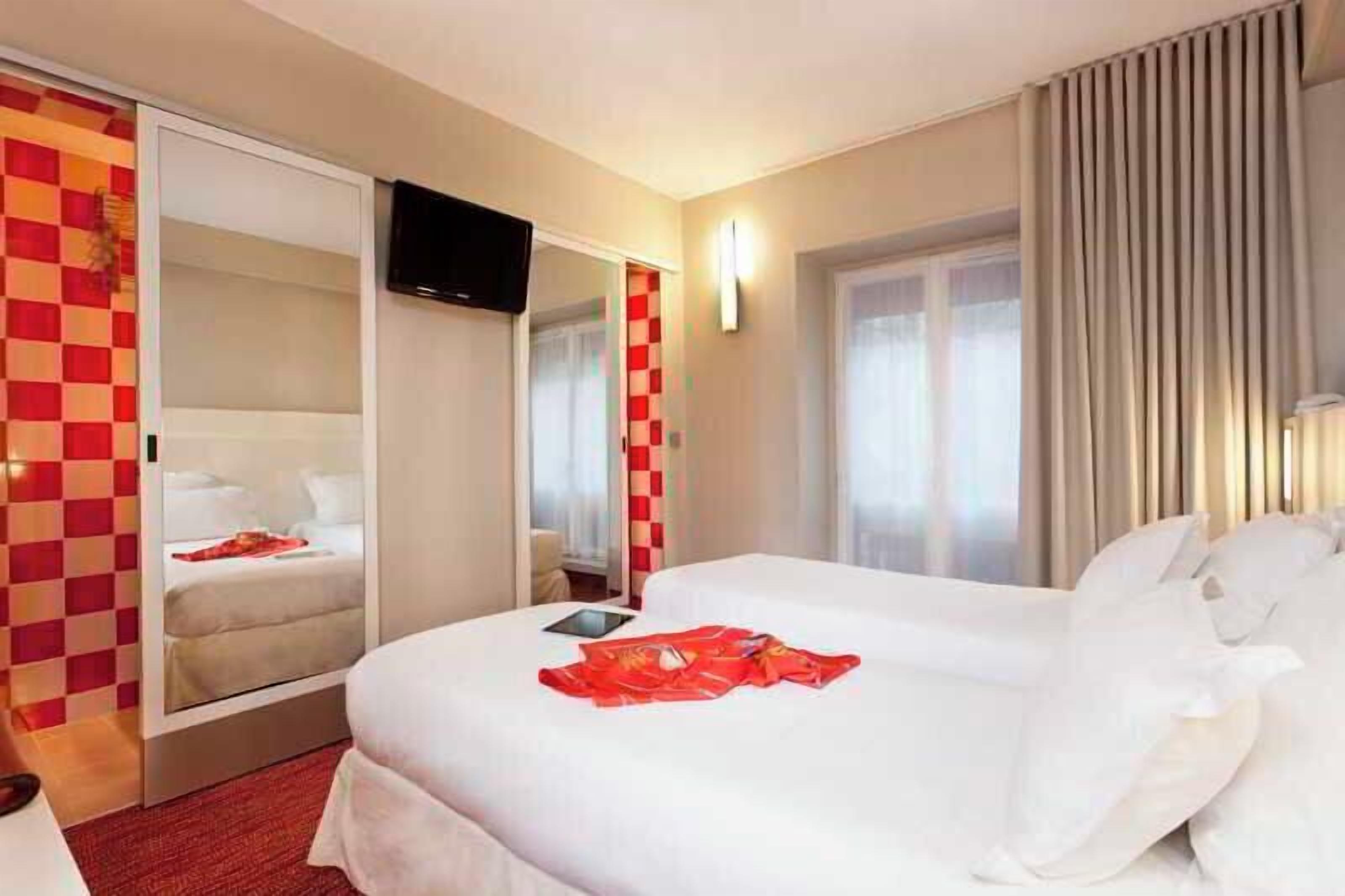 Una camera d'albergo con un letto, una televisione, uno specchio e una finestra con tende.