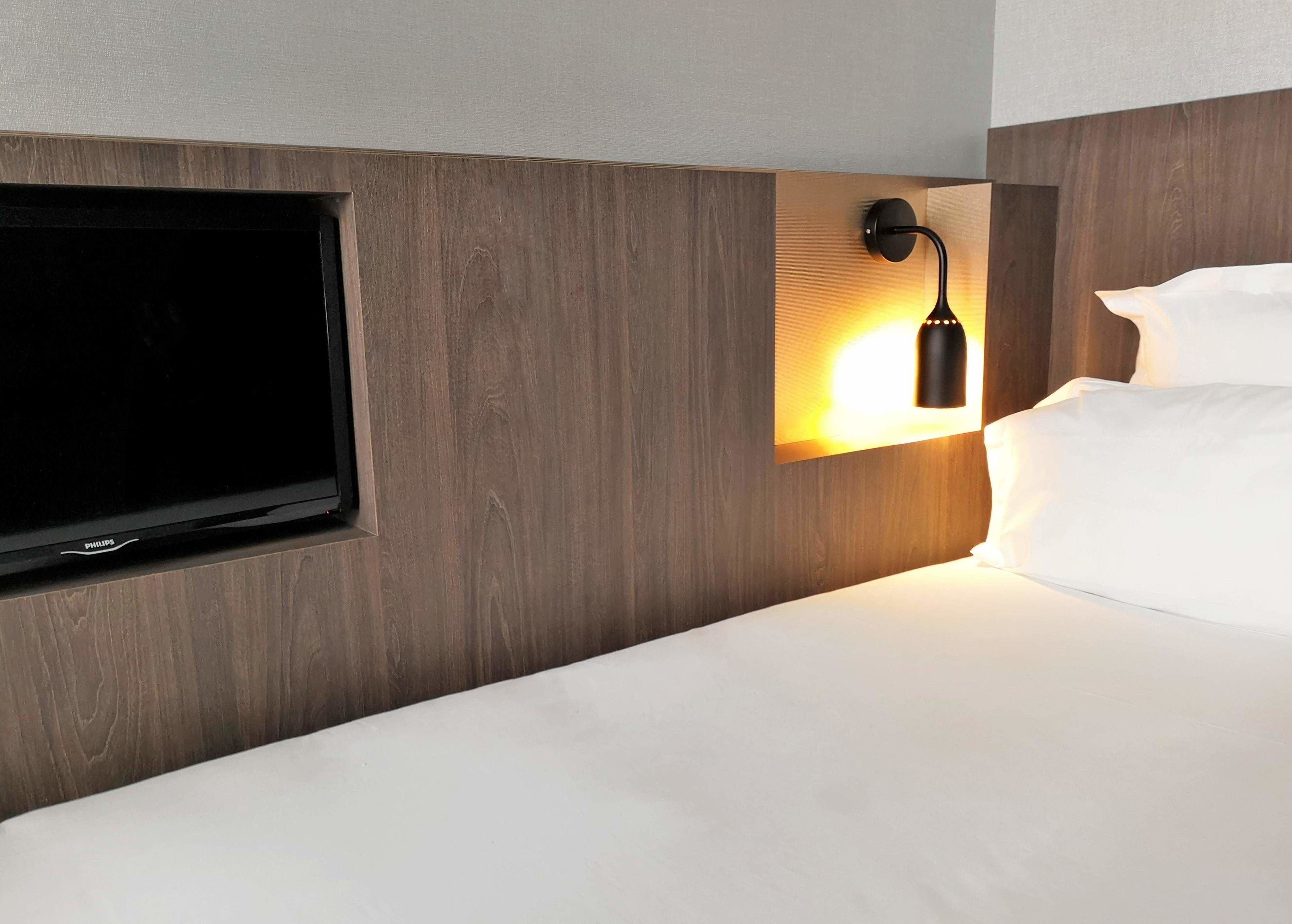 A hotel room with a flat-screen TV mounted on a wooden panel wall, a bedside lamp, and a neatly made bed with white linens.