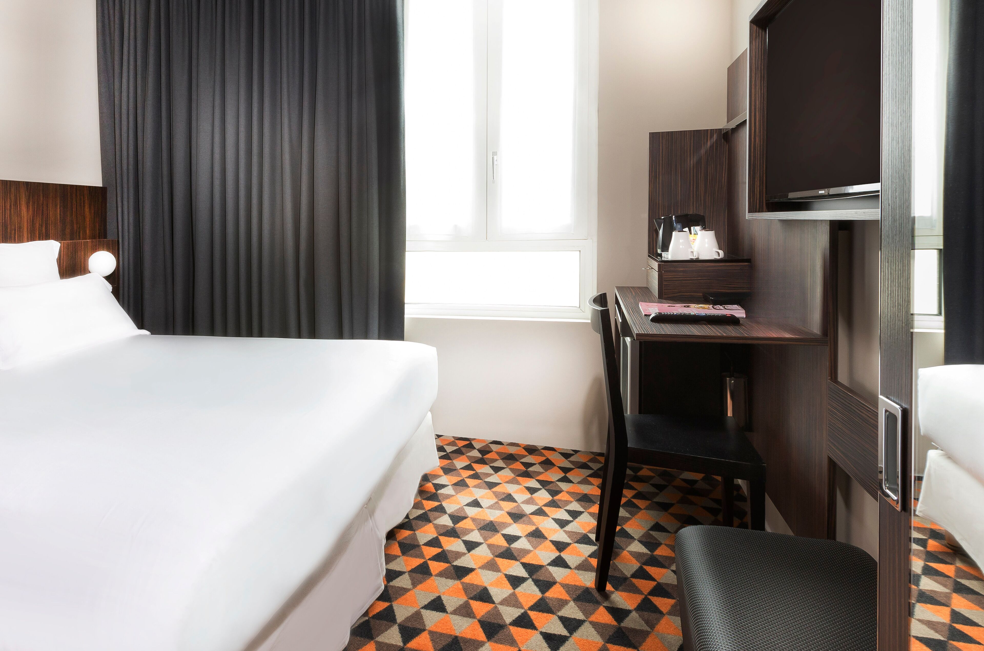 Une chambre d’hôtel comprenant un lit, un bureau avec une chaise, une télévision et une fenêtre avec des rideaux.