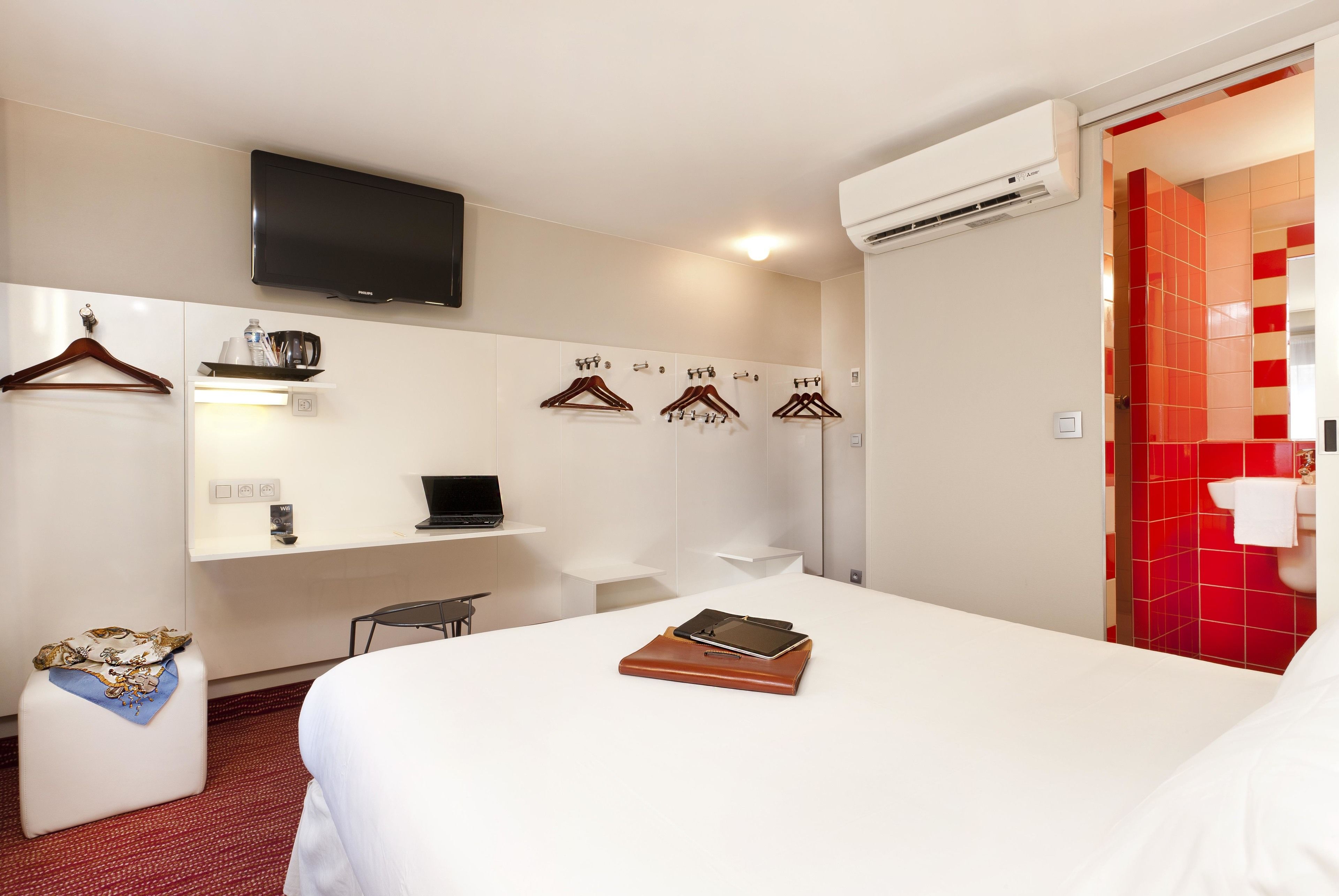 A hotel room with a bed, a desk with a laptop, a TV mounted on the wall, and a red-tiled shower area.
