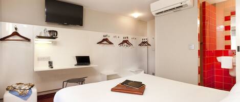 A hotel room with a bed, a desk with a laptop, a TV mounted on the wall, and a red-tiled shower area.