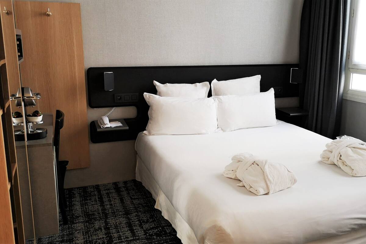 Hotelkamer met een bed, nachtkastje met telefoon en lamp, een houten kledingkast en een raam met gordijnen.