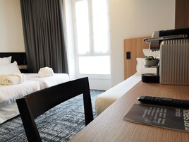 A hotel room with a bed, a desk with a remote control, a coffee machine, and a window with curtains.