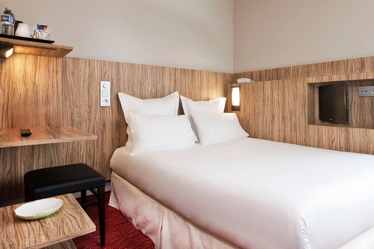 Hotelkamer met een houten wandpaneel, een bed met witte beddengoed, een nachtkastje met een schaal en een klein plankje met toiletartikelen.