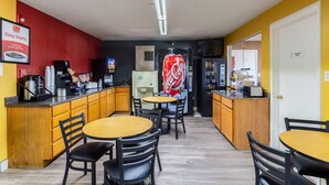 Uma cozinha compacta com armários de madeira, uma máquina de café, uma geladeira Coca-Cola e um micro-ondas. Há duas mesas com cadeiras.