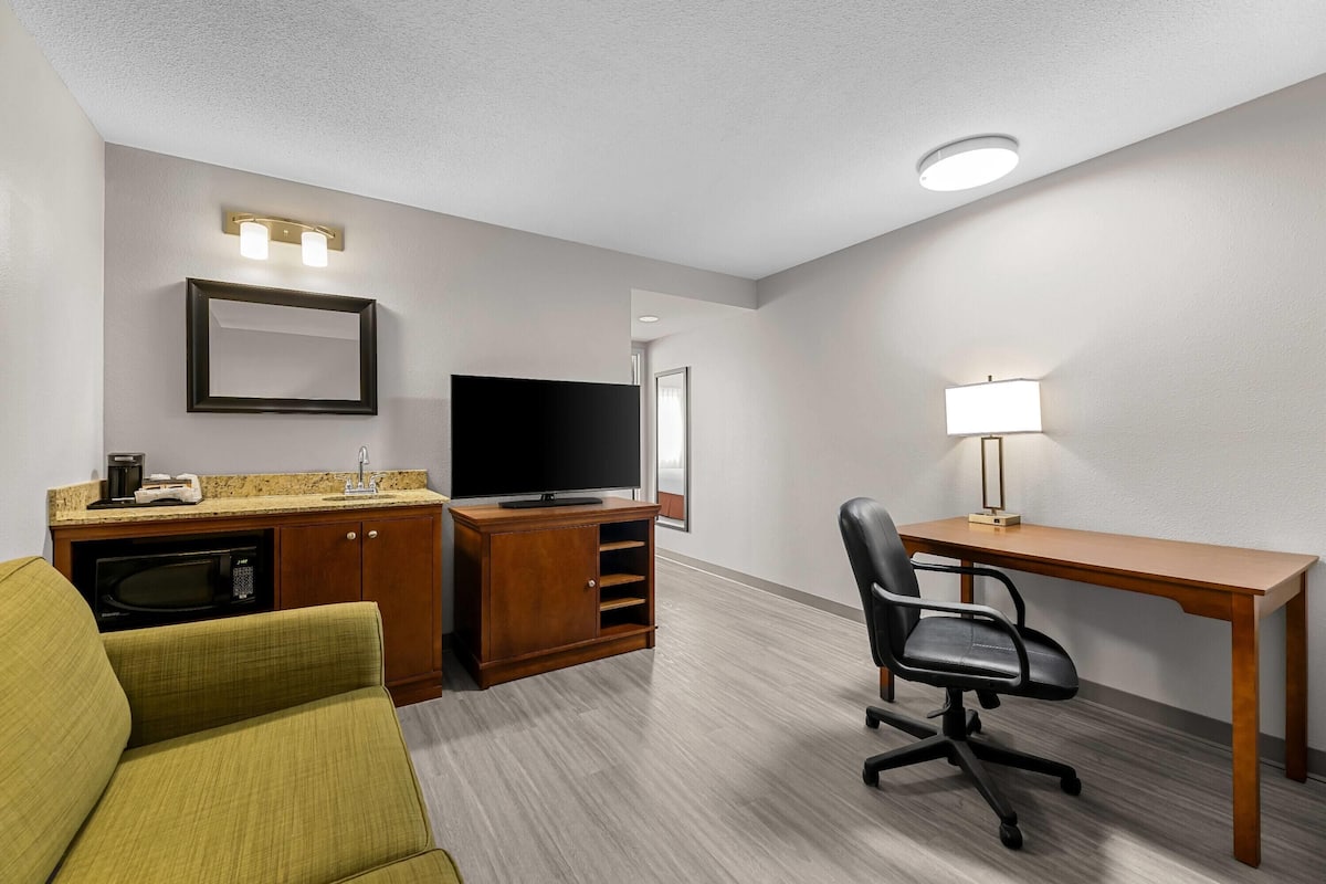 A hotel room with a kitchenette, a television, a desk with a lamp, a chair, and a sofa.