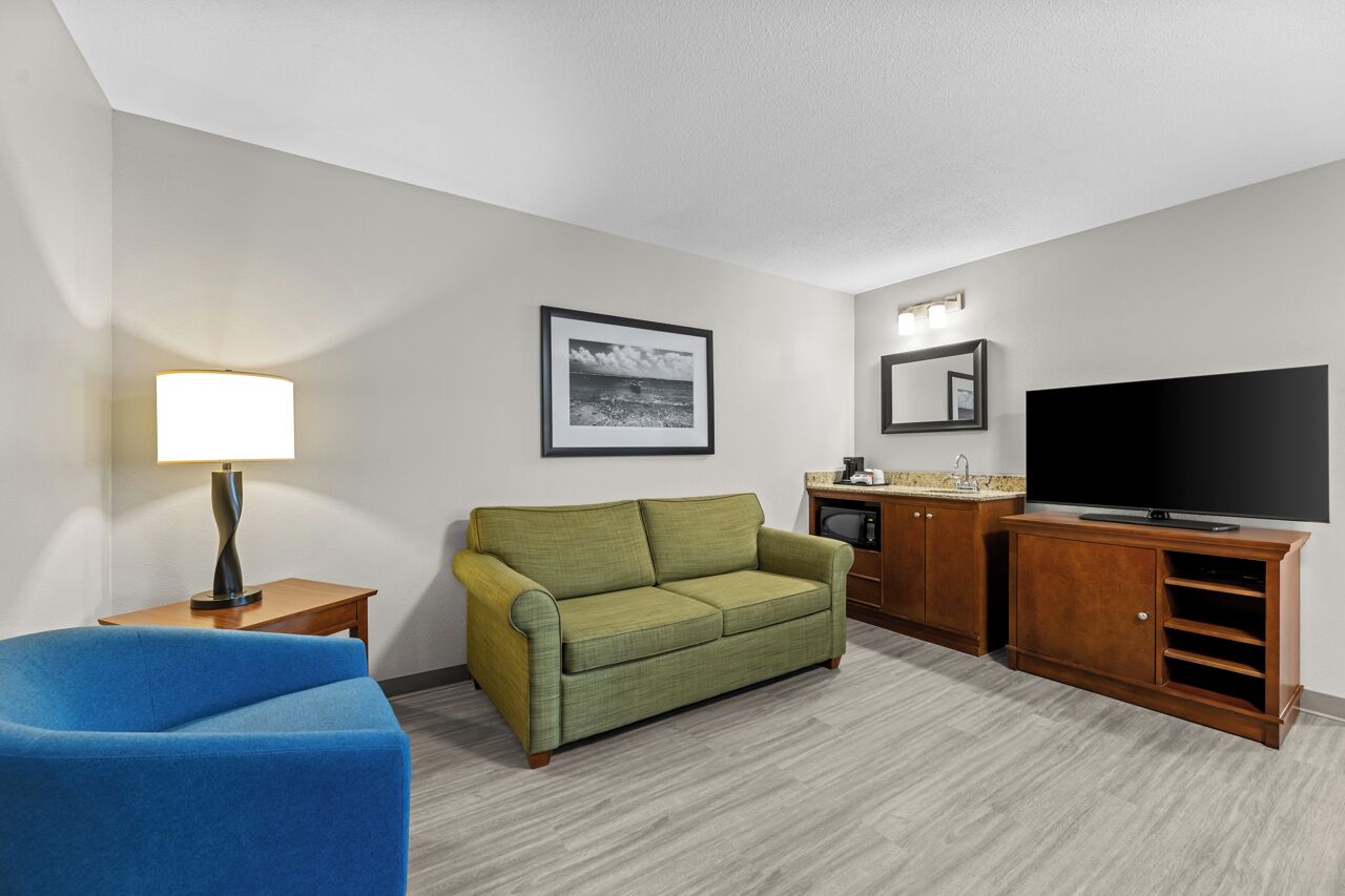 A hotel room with a green sofa, a blue armchair, a TV, a small kitchenette, and a framed picture on the wall.