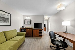 Une pièce de taille modeste, avec un canapé vert, un bureau en bois, un fauteuil de bureau noir, une télévision et une lampe.