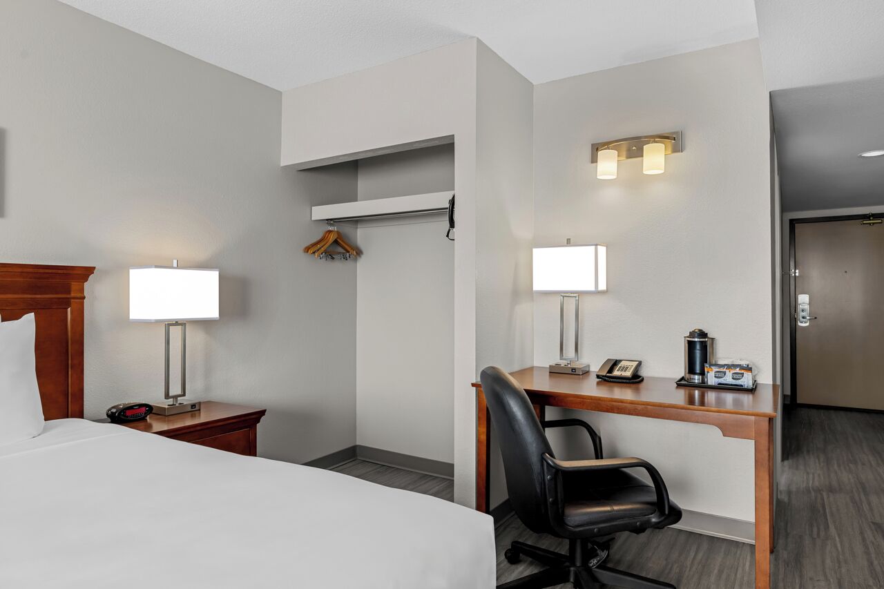 Una habitación de hotel con cama, mesita de noche, lámpara, escritorio, silla y perchero.