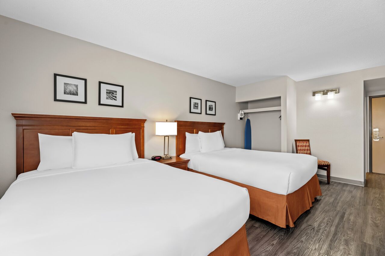 A hotel room with two beds, a nightstand with a lamp, a chair, and a coat rack.