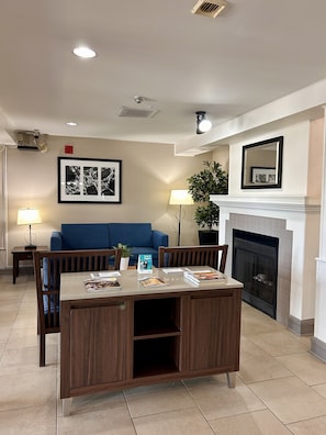 A living room with a fireplace, a dining table with chairs, a blue sofa, and a lamp. - Travelodge Suites by Wyndham Regina / Eastgate Bay (Regina)