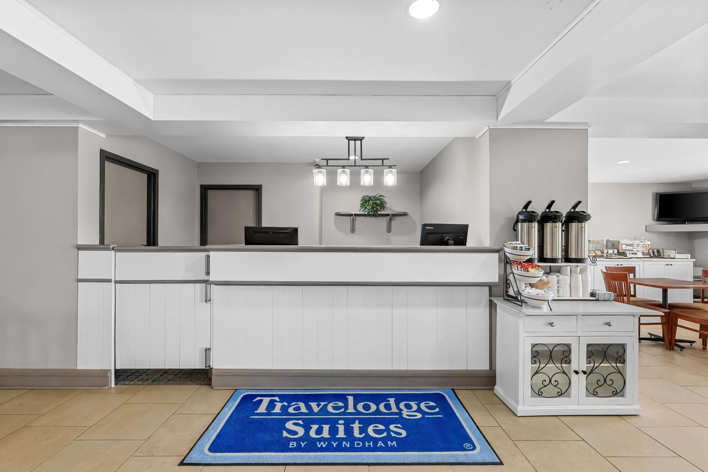 Eine Küchenzeile mit weißen Schränken, einer blauen Matte mit der Aufschrift 'Travelodge Suites by Wyndham', einer kleinen Pflanze und einem Fernseher.