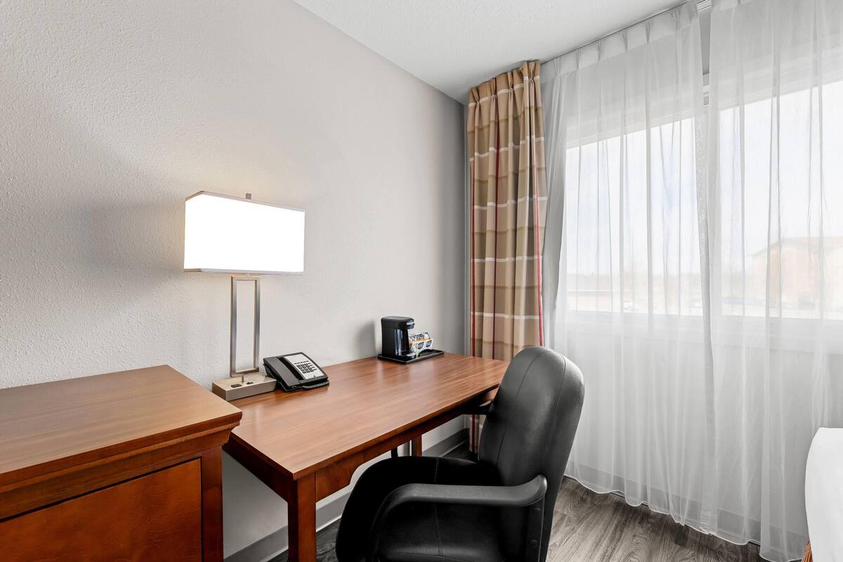 A hotel room with a wooden desk, a black leather chair, a lamp, a phone, and a coffee maker.
