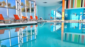 An indoor swimming pool with clear blue water, orange lounge chairs, and a modern design with large windows and a blue wall. - Hotel Huntington Beach (Huntington Beach)
