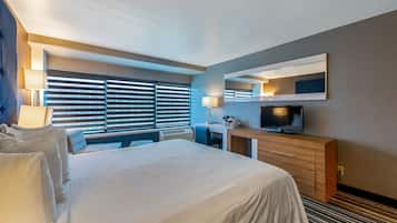 A modern hotel room with a large bed, a desk, a television, and a window with blinds.