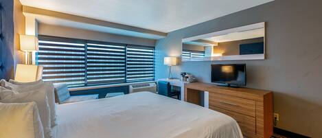 A modern hotel room with a large bed, a desk, a television, and a window with blinds.