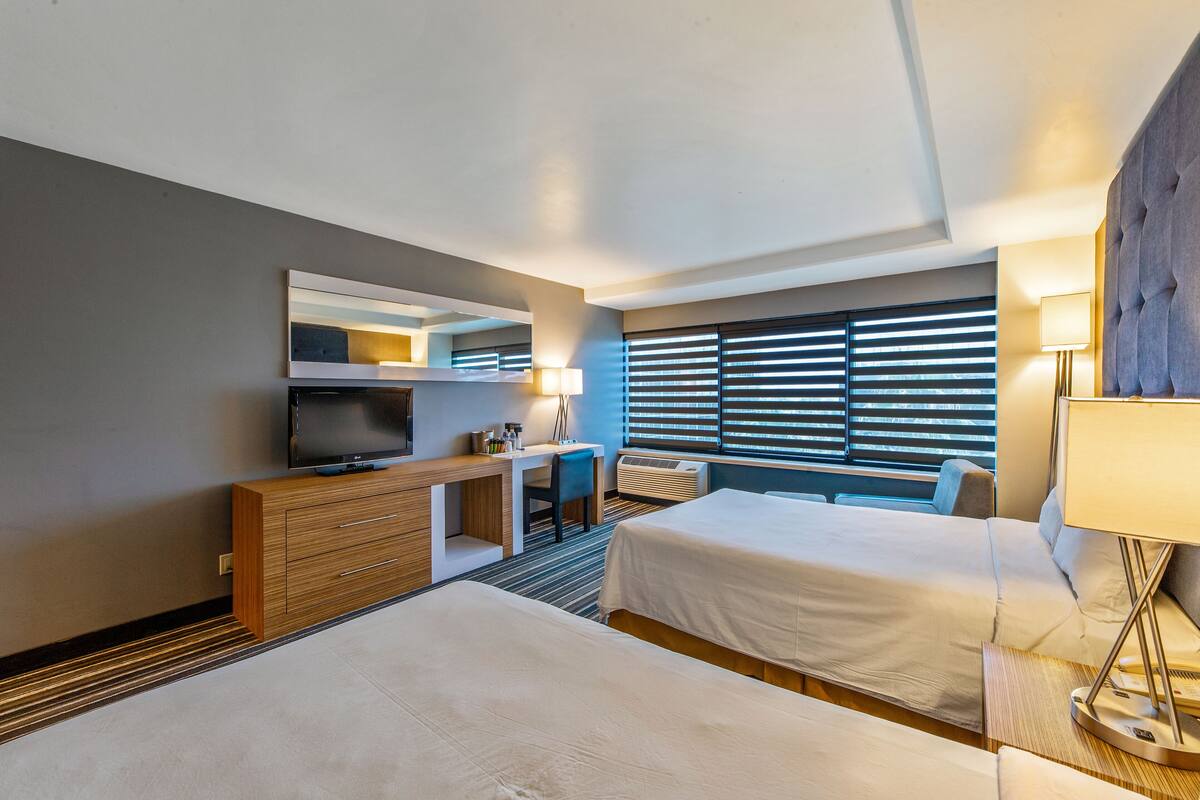 A modern hotel room with two beds, a desk, a television, and a large window with blinds.