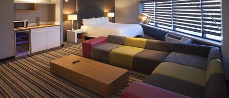 Habitación de hotel con cama, sofá, mesa de centro, escritorio con microondas y ventana con persianas.