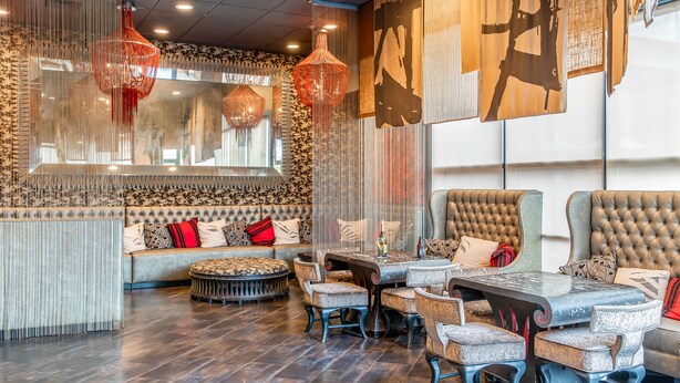 A restaurant interior with a high ceiling, hanging red pendant lights, a large mirror, and a seating area with a sofa and cushioned chairs. - Hotel Huntington Beach (Huntington Beach)