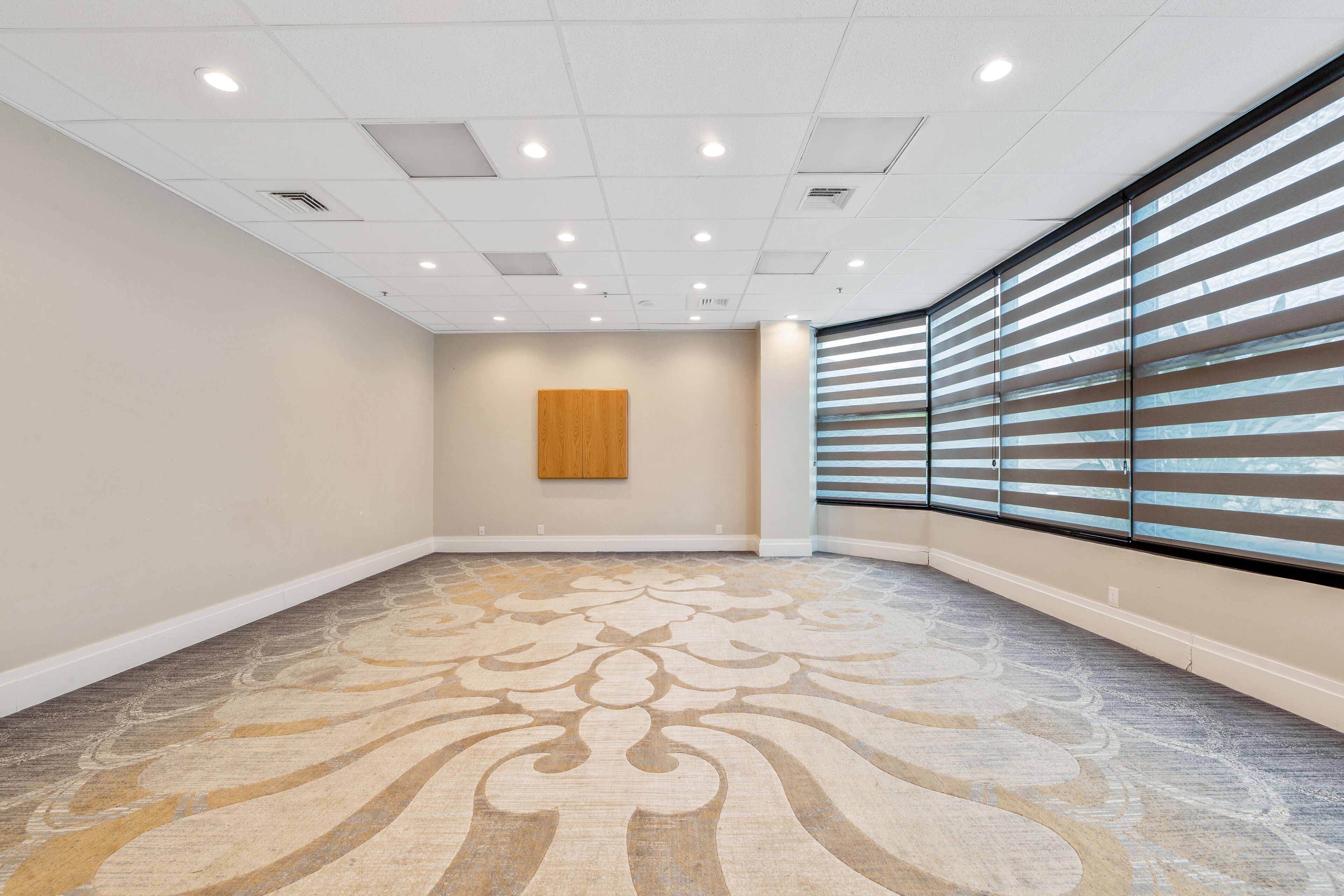 Una habitación amplia y luminosa con alfombra estampada, un panel de madera decorativo y ventanas grandes con persianas.