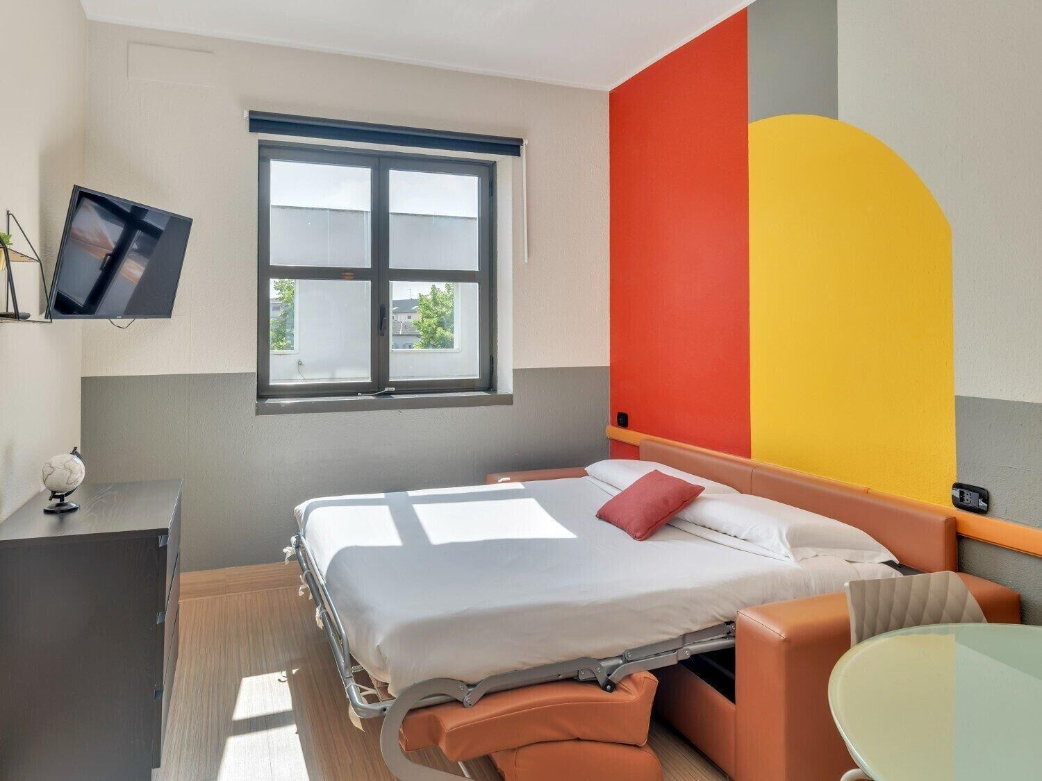 A hotel room with a bed, a desk, a chair, a TV, and a window with a view of buildings.