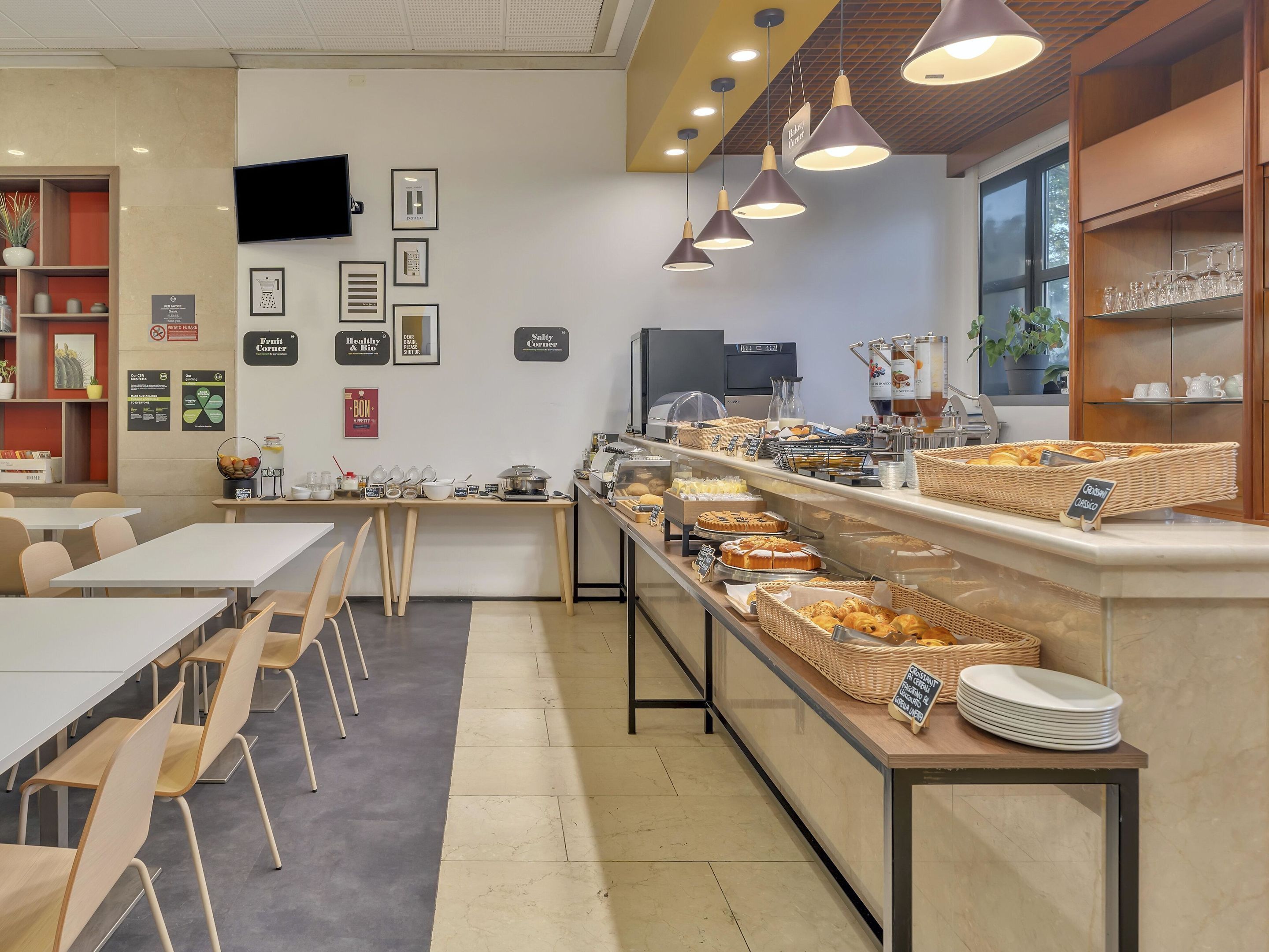 Un café con una barra larga que exhibe pasteles y café, mesas con sillas y un televisor montado en la pared.
