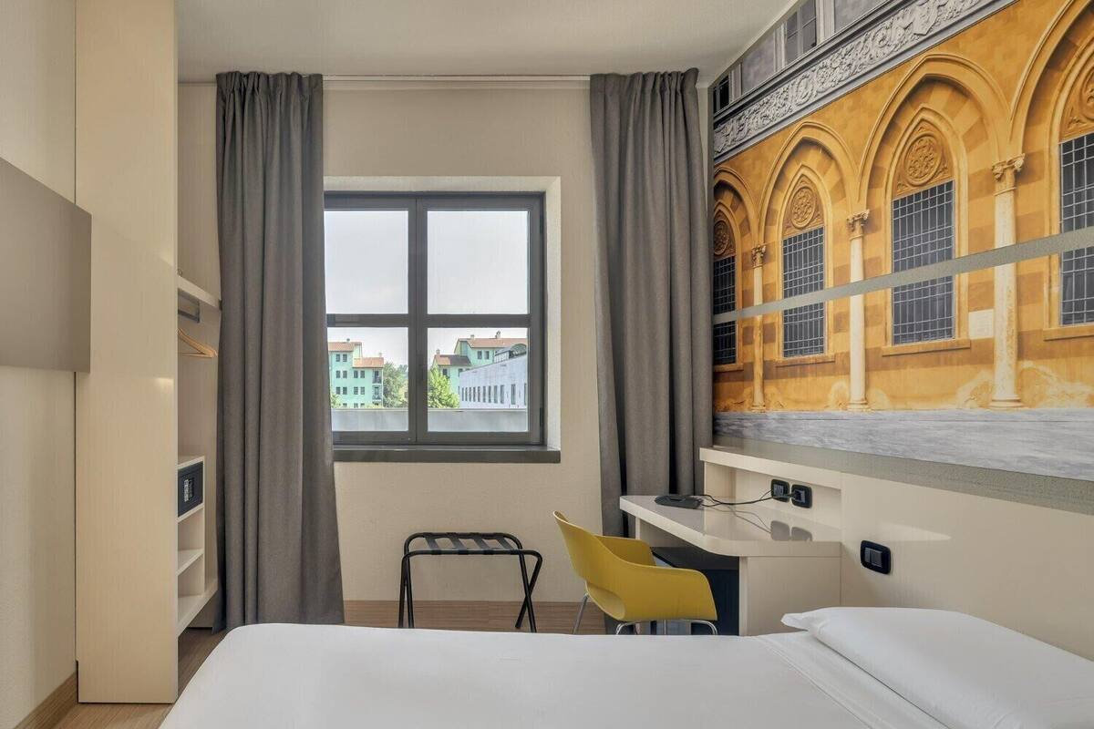A hotel room with a bed, a desk with a chair, a window with curtains, and a mural on the wall.
