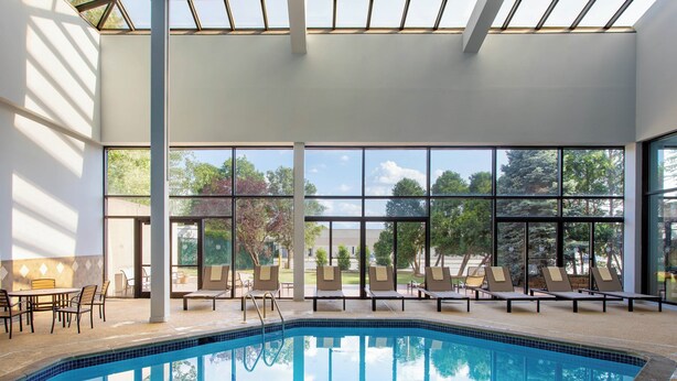 Una piscina interior con paredes de vidrio transparente, sillas de descanso y una mesa.