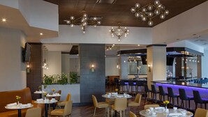 Un comedor moderno con mesas y sillas redondas, una zona de bar con barra iluminada en azul, y una planta grande en el centro.